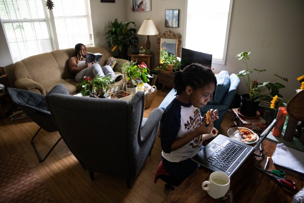 A girl eats in front of her laptop while her mother reads in the background
