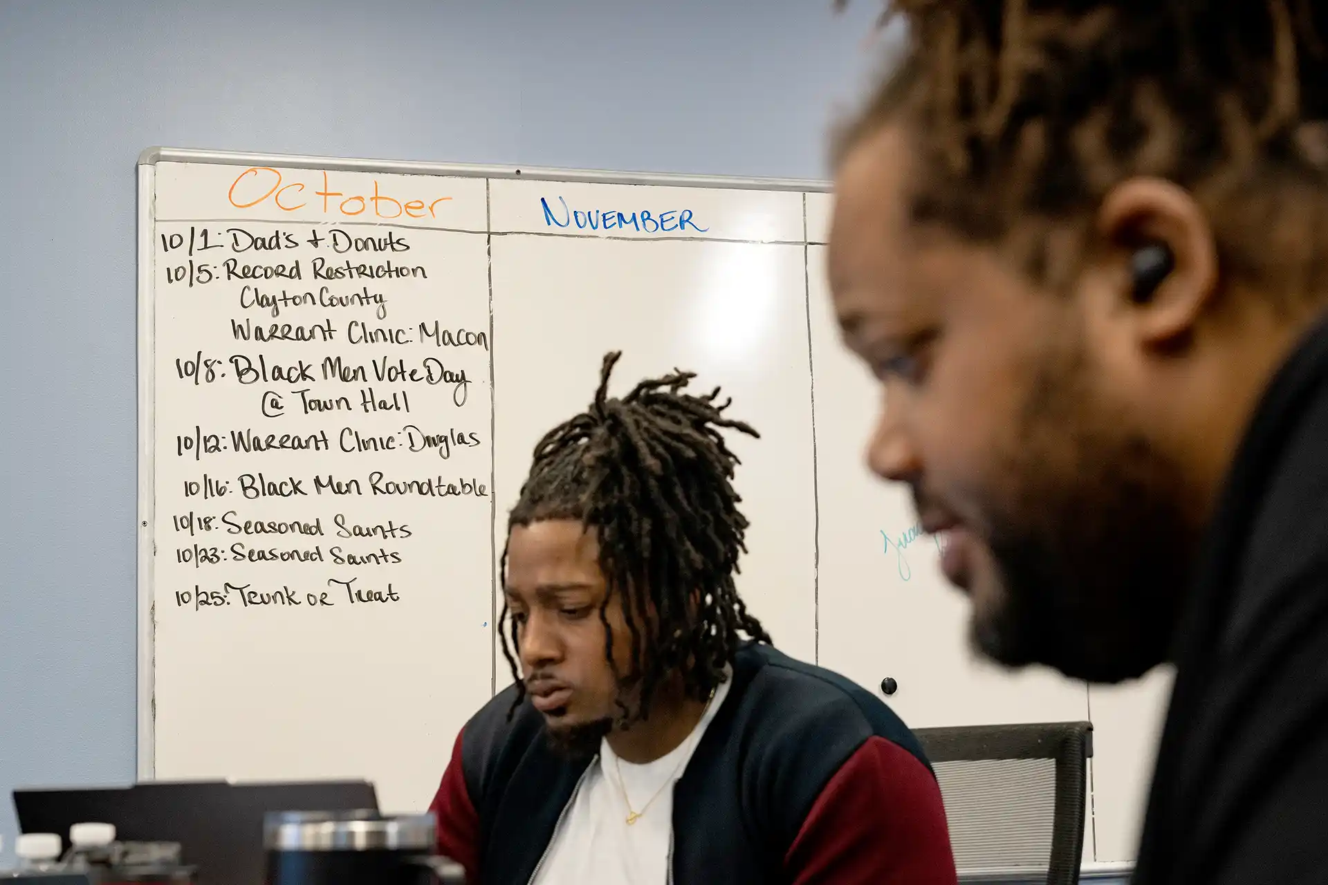 Two men sit at a table in front of a whiteboard.