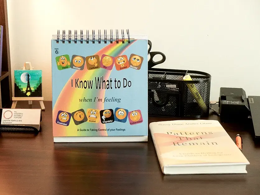 desktop-flipchart-trauma-book A psychotherapist's desk features an 'I Know What to Do When I'm Feeling' flip book and a copy of 'Patterns That Remain: A Guide to Healing for Asian Children of Immigrants.'